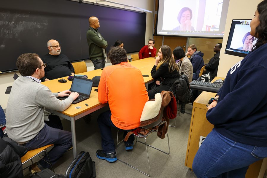A group of individuals sit around a table in a classroom attending the breakout session focused on general discussion of computer vision, multimodality, and search-related abstracts. A projector screen at the front of the room depicts virtual participants.